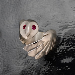 May include: A silver owl ring with ruby-colored eyes. The ring features detailed wings and a textured finish, creating a unique and artistic design. The owl's face is centered, with the wings extending on either side.