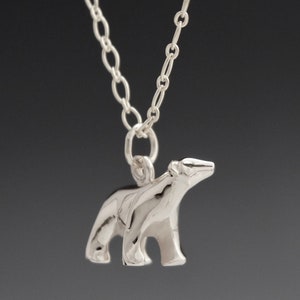 May include: A silver-colored necklace featuring a pendant in the shape of a polar bear. The bear is standing on all fours, with its head raised. The chain is made of small, linked circles. The background is a dark gray.