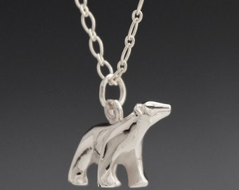 May include: A silver-colored necklace featuring a pendant in the shape of a polar bear. The bear is standing on all fours, with its head raised. The chain is made of small, linked circles. The background is a dark gray.