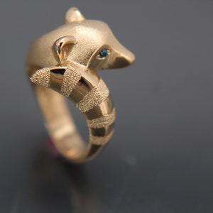 May include: A gold-colored ring featuring a raccoon design. The raccoon's head is at the top, with a textured band forming the ring's base. The raccoon has a small blue gem for an eye. The ring is set against a dark background.