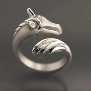 May include: A silver-colored ring in the shape of a unicorn's head. The ring wraps around the finger with the unicorn's head on top and a detailed mane and horn. The ring is made of a shiny metal and is set against a dark background.