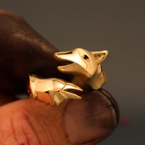 Könnte beinhalten: Ein goldfarbener Ring mit einem Fuchsmotiv. Der Ring umschließt den Finger mit entgegengesetzten Fuchsköpfen. Die Fuchsköpfe weisen detaillierte Merkmale auf, darunter spitze Ohren und offene Mäuler.