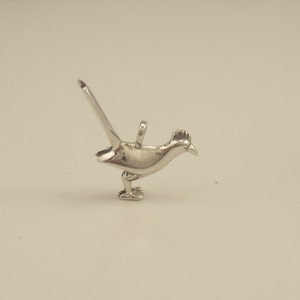 May include: A polished silver pendant in the shape of a roadrunner bird. The bird is depicted in a running pose with its tail extended. The pendant has a loop for attaching to a chain or bracelet. The roadrunner has a detailed crest and beak.