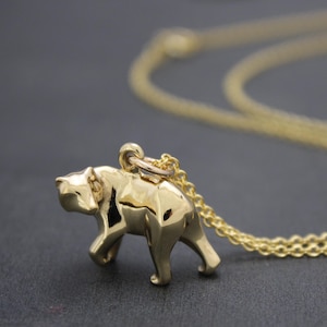 May include: A gold-colored bear pendant necklace. The bear is depicted in a geometric, angular style, with a polished finish. The necklace chain is also gold and is visible against a dark background. The bear is walking.