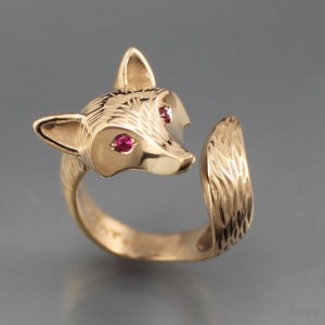 May include: A gold-colored fox ring with ruby-colored eyes. The fox head is detailed with etched fur and the tail wraps around the finger. The ring is a unique piece of jewelry.