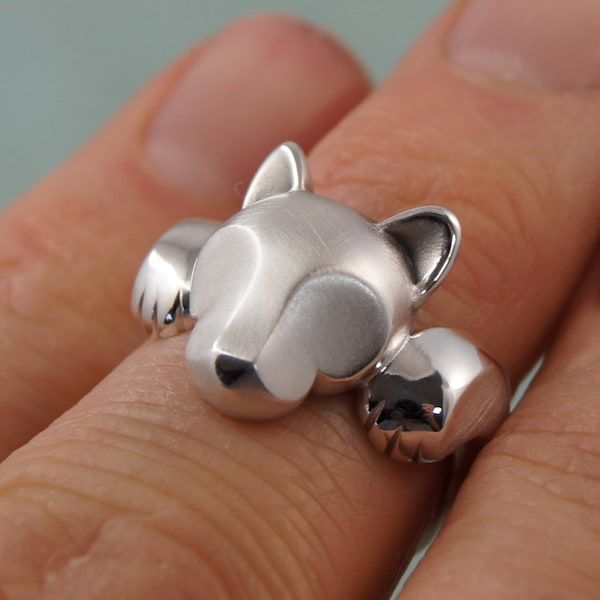 May include: A silver ring featuring a stylized cat design. The ring has a cat's head and paws, with the head positioned on top of the finger and the paws on either side. The ring is made of a shiny, metallic material.