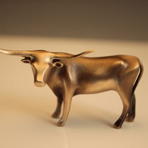 Puede incluir: Una escultura de color bronce de un toro Longhorn. El toro est&aacute; de pie con la cabeza ligeramente levantada, mostrando sus largos cuernos curvados. La escultura tiene un acabado liso y pulido, y los detalles de los m&uacute;sculos del toro son visibles.