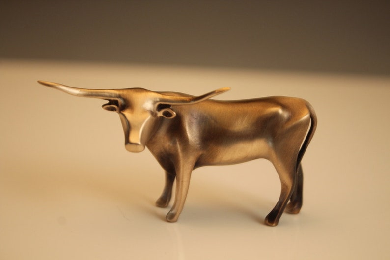 Puede incluir: Una escultura de color bronce de un toro Longhorn. El toro est&aacute; de pie con la cabeza ligeramente levantada, mostrando sus largos cuernos curvados. La escultura tiene un acabado liso y pulido, y los detalles de los m&uacute;sculos del toro son visibles.