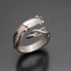 May include: A silver-colored ring in the shape of a squid. The ring features two small red gemstones and detailed tentacles. The design is open, allowing for an adjustable fit. The ring is set against a dark gray background.