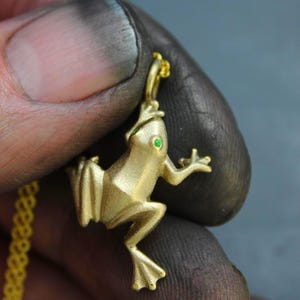 May include: A gold-colored frog pendant with a green gemstone eye. The frog is detailed with textured surfaces and is suspended from a delicate gold chain. The pendant is held by a hand with dark smudges.