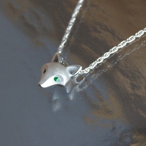 May include: A silver fox pendant necklace. The pendant is a stylized fox head with green gemstone eyes. The necklace chain is a delicate silver rope. The pendant and chain are displayed on a reflective surface.