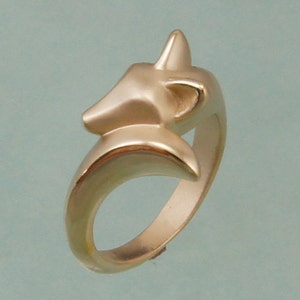 May include: A gold-colored ring featuring a stylized fox head design. The ring's band curves smoothly, with the fox's head positioned at the top, showcasing its ears and snout. The ring is a piece of jewelry.