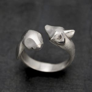 May include: A silver ring with two animal head designs facing each other. One head has pointed ears and an open mouth, the other is smooth and rounded. The ring is set against a dark background.