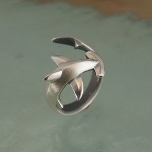 May include: A silver-colored ring featuring a hammerhead shark design. The ring has a detailed, three-dimensional shark with a textured finish. The shark's head and body wrap around the band, creating a unique and eye-catching piece of jewelry.