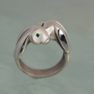 May include: A silver ring featuring two stylized rabbits embracing. Each rabbit has a green gemstone eye. The ring has a brushed metal finish and a smooth, rounded band.