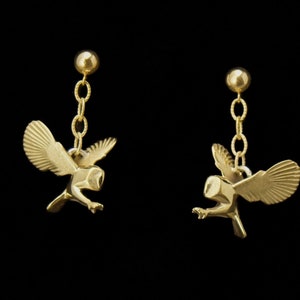 May include: A pair of gold-colored owl earrings. Each earring features a small, detailed owl pendant with outstretched wings, suspended from a chain and a round stud. The earrings are set against a black background.