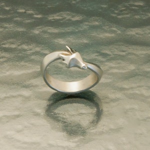 May include: A silver ring featuring a stylized fox head design. The ring has a smooth, matte finish and a small, clear gemstone accent. The fox head is positioned on the top of the ring, with the band curving around the finger. A unique piece of jewelry.