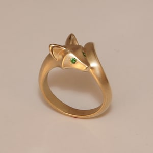 May include: A gold-colored ring in the shape of a fox. The fox's eyes are small green gemstones. The ring has a smooth, matte finish. The fox's head is turned to the side, and the tail wraps around the band.