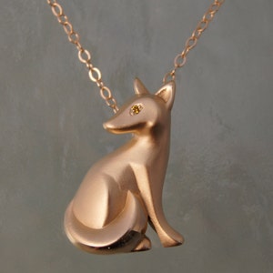 Puede incluir: Un collar con colgante de zorro en oro rosa. El zorro está sentado con la cola enroscada y tiene una pequeña gema amarilla como ojo. La cadena también es de oro rosa.