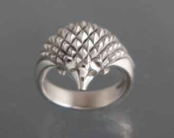 May include: A silver ring featuring a hedgehog design. The ring's band is smooth, leading to a detailed hedgehog with textured quills. The hedgehog's face has small, defined features. The ring is likely made of sterling silver.