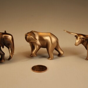 Puede incluir: Tres esculturas de animales de bronce: un elefante, un oso y un toro. El elefante y el oso caminan, mientras que el toro est&aacute; de pie con la cabeza levantada. Una moneda est&aacute; en el suelo frente a las esculturas.