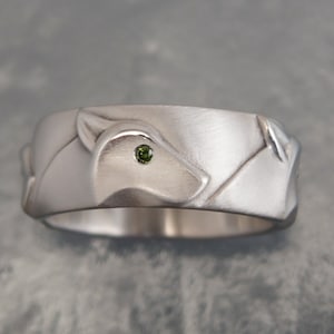 May include: A silver ring with a fox design. The ring features a detailed fox head with a small green gemstone eye. The band has a textured, matte finish. The ring is a piece of jewelry.