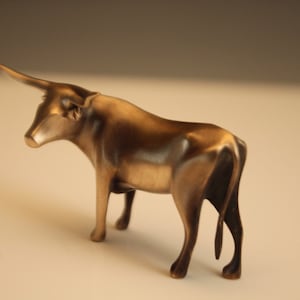 Puede incluir: Escultura de bronce de un toro con un solo cuerno. El toro est&aacute; representado de pie, con la cabeza girada hacia la izquierda. La escultura tiene un acabado met&aacute;lico pulido y se encuentra sobre un fondo neutro. La forma del toro es suave y detallada.