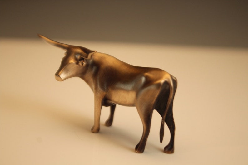 Puede incluir: Escultura de bronce de un toro con un solo cuerno. El toro est&aacute; representado de pie, con la cabeza girada hacia la izquierda. La escultura tiene un acabado met&aacute;lico pulido y se encuentra sobre un fondo neutro. La forma del toro es suave y detallada.