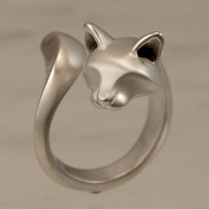 May include: A silver-toned ring featuring a fox design. The ring wraps around the finger with the fox's head at the top and the tail curving around the side. The fox's face has detailed features, including pointed ears and a defined snout.