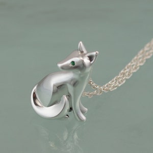 May include: A silver fox pendant necklace. The fox is in a seated position with a curved tail and green eyes. The necklace chain is a delicate silver. The pendant is highly polished and reflects the light.