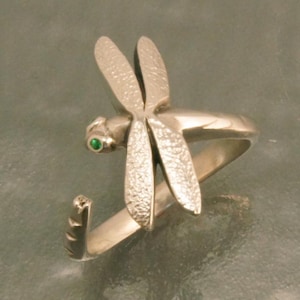 May include: A silver dragonfly ring with textured wings and a green gemstone eye. The ring features a detailed dragonfly design, with the wings spread and the body curving around the finger. The ring is made of a shiny metal.