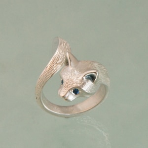 May include: A silver-toned ring featuring a fox design. The fox's head is detailed with blue gemstone eyes and textured fur. The tail curves around the finger, creating a unique and whimsical piece of jewelry.