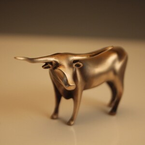 Puede incluir: Figurilla de toro de color bronce con cuernos largos y cuerpo detallado. La escultura est&aacute; de pie sobre cuatro patas, con un acabado liso y pulido. El toro es un objeto decorativo.