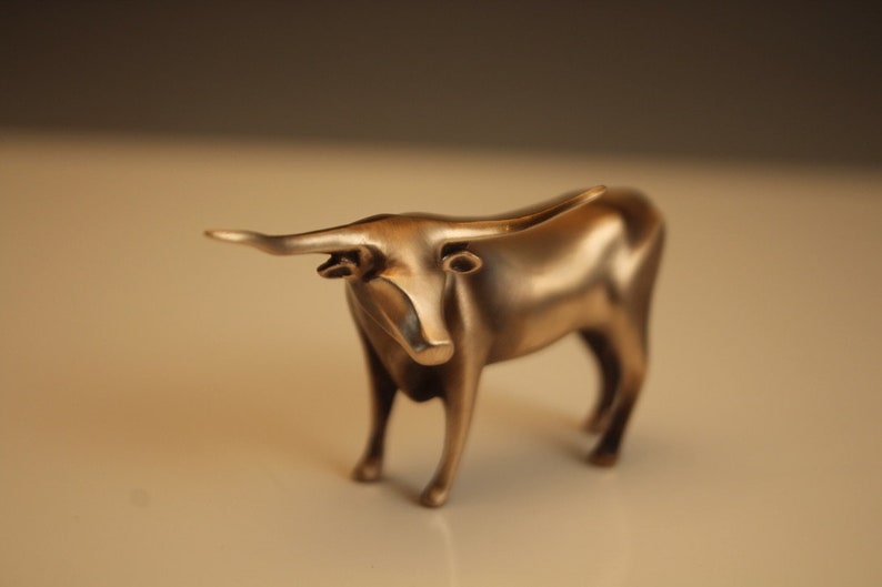 Puede incluir: Figurilla de toro de color bronce con cuernos largos y cuerpo detallado. La escultura est&aacute; de pie sobre cuatro patas, con un acabado liso y pulido. El toro es un objeto decorativo.