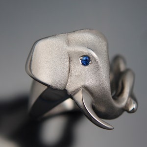 May include: A silver elephant ring with a textured finish. The elephant's eye features a small, round, blue gemstone. The trunk is curled, and the tusks are visible. The ring is set against a blurred background.