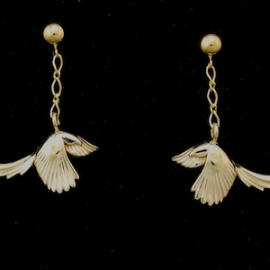 May include: A pair of gold-colored bird earrings. Each earring features a small, detailed bird pendant with outstretched wings, suspended from a chain and a round stud. The earrings are set against a black background.