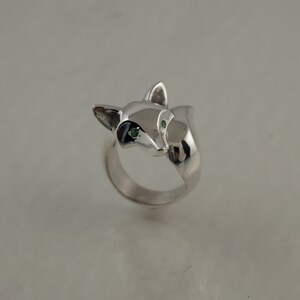 May include: A silver-colored ring featuring a fox head design. The fox has green eyes and black accents around the eyes. The ring is a solid, polished metal with a smooth finish. The fox's head is the focal point of the ring.