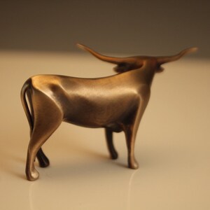 Puede incluir: Escultura de bronce de un toro con largos cuernos. El toro est&aacute; de pie con la cabeza ligeramente girada. La escultura tiene un acabado pulido y se encuentra sobre un fondo neutro. Mide aproximadamente 20 cm de largo.