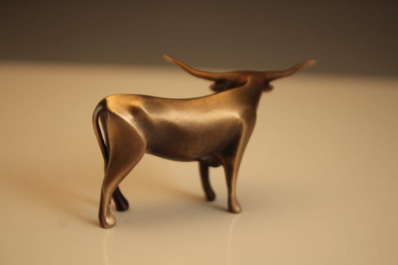 Puede incluir: Escultura de bronce de un toro con largos cuernos. El toro est&aacute; de pie con la cabeza ligeramente girada. La escultura tiene un acabado pulido y se encuentra sobre un fondo neutro. Mide aproximadamente 20 cm de largo.