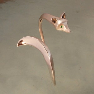 May include: A silver-toned fox-shaped sculpture with a curved tail and a slender neck. The fox's head has green gemstone eyes. The sculpture is a decorative item, possibly a piece of jewelry or home decor.