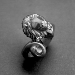 May include: A silver-toned ring featuring a detailed ram's head design. The ram's head has textured fur and a prominent nose. The ring's band curves into a spiral shape. The ring is likely made of metal and is a piece of jewelry.