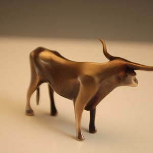 Puede incluir: Figura de toro de color bronce, con cuernos curvados y una constituci&oacute;n musculosa. La escultura est&aacute; de pie sobre cuatro patas, con la cabeza ligeramente girada hacia un lado. Es un objeto decorativo.