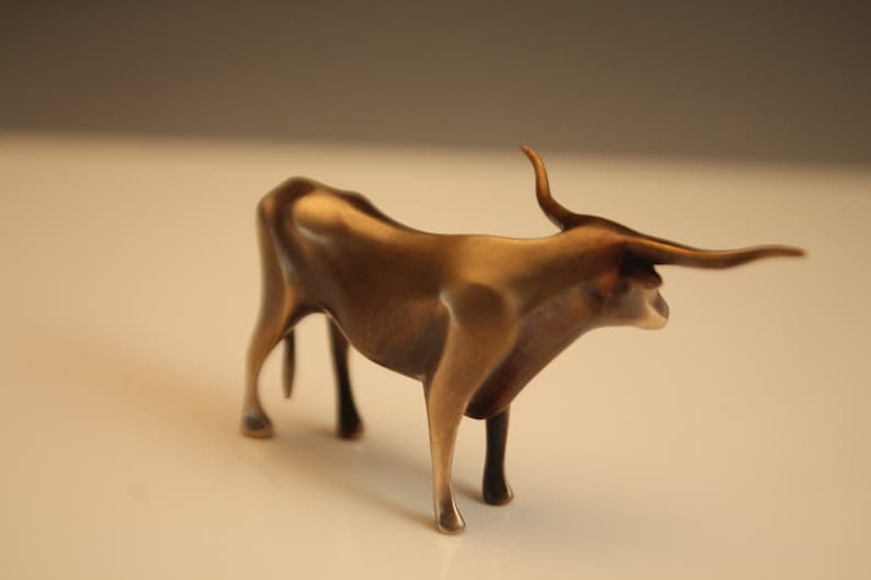 Puede incluir: Figura de toro de color bronce, con cuernos curvados y una constituci&oacute;n musculosa. La escultura est&aacute; de pie sobre cuatro patas, con la cabeza ligeramente girada hacia un lado. Es un objeto decorativo.