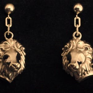 May include: A pair of gold-colored lion head dangle earrings. Each earring features a detailed lion's head pendant suspended from a chain and a small ball stud. The earrings are set against a dark background.
