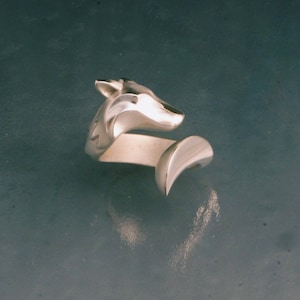May include: A silver-toned ring featuring a wolf head design. The ring wraps around the finger with a curved, open-ended band. The wolf's head is detailed with carved features, and the ring has a polished finish.