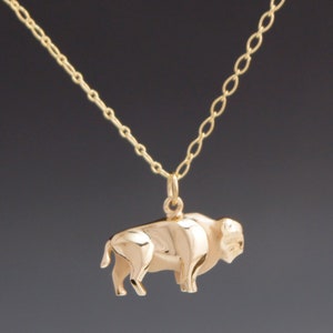 May include: A gold-colored necklace featuring a pendant in the shape of a bison. The bison pendant is detailed and hangs from a delicate chain. The necklace is displayed against a dark background.