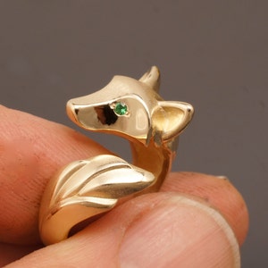 May include: A gold-colored fox ring with a green gemstone eye. The ring features a detailed fox head and tail design. The ring is shown in a close-up shot, highlighting the craftsmanship and the gemstone's sparkle.