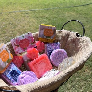 May include: A basket filled with various handmade soaps. The soaps are in different shapes and colors, including pink, purple, and yellow. Some soaps are floral-shaped, and others are rectangular. The basket has a burlap lining and a metal handle. The words "Handmade All Natural" are visible on some of the soap packaging.