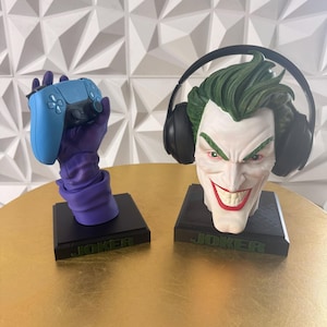 May include: Two gaming-themed statues. One features a blue game controller held by a purple gloved hand. The other is a bust of the Joker with green hair and headphones. Both are on black bases with "JOKER" in green.