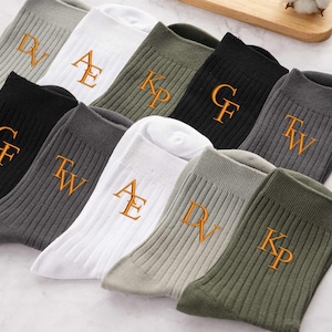 May include: Assorted ribbed crew socks in various colors, including gray, white, black, and olive green. Each sock features embroidered initials in a contrasting gold color. The socks are neatly arranged, showcasing the personalized designs.